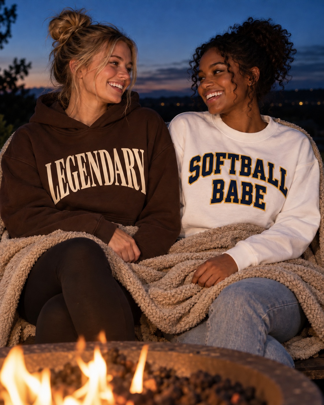 two friends sharing a blanket beside a fire pit at dusk, deep blue sky behind them, close and candid