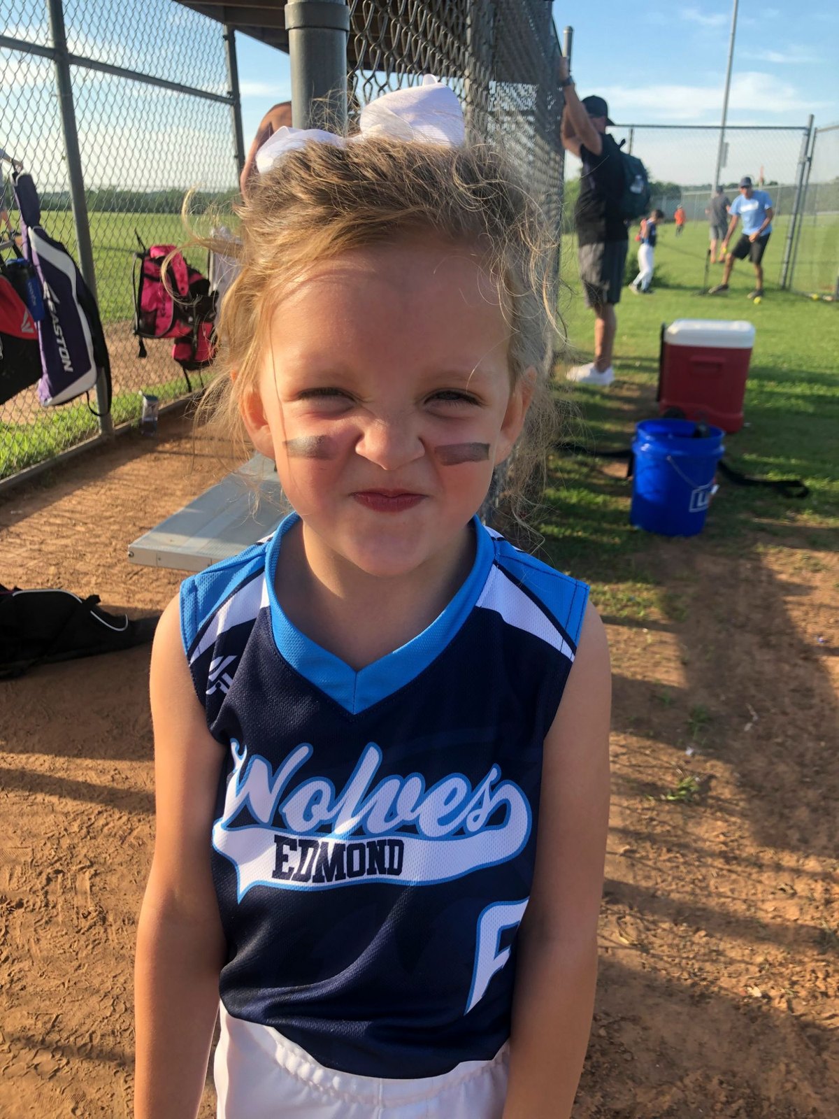 Genevieve in her first softball season, in a Wolves jersey and eye black by the dugout fence